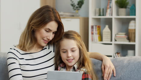 close up of the beautiful mother sitting on the sofa and hugging her pretty teen daughter while they watching something on the tablet device