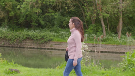 Woman-Exercising-Outdoors-Walking-Along-Bank-Of-River-With-Duck-In-Background