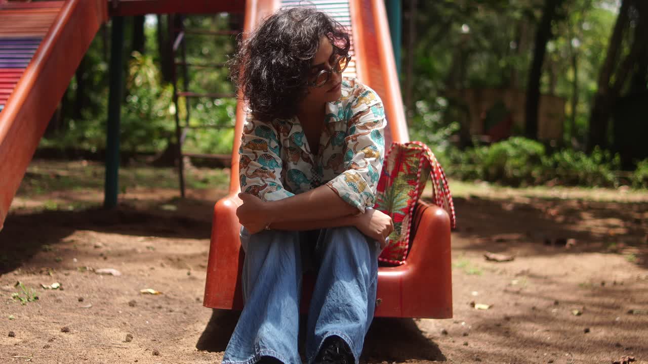 A woman in casual outfit sits at the bottom of a playground slide with a colorful bag on the side