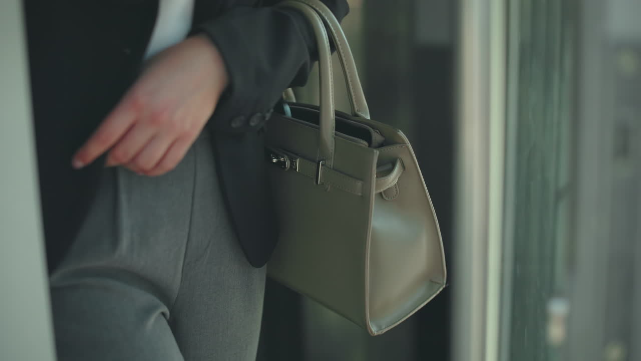 ハンドバッグを持ち、黒いジャケットとグレーのズボンという профессиона attireを身につけ、モールから出てくる女性の部分的なクローズアップビュー。ガラスのドアが周囲の光を反射しています。