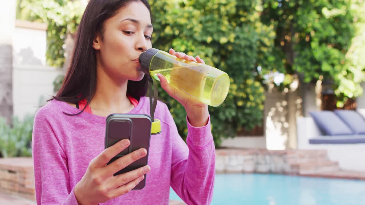 mujer biracial usando un teléfono inteligente y bebiendo agua en el jardín