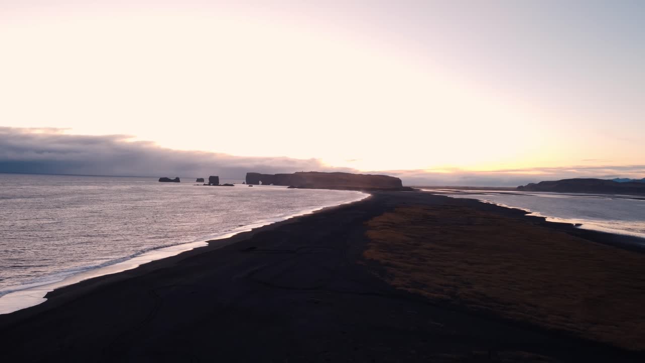 A tranquil black sand beach at Reynisfjara, Iceland, stretches into the distance, reflecting the soft hues of a sunset or sunrise over the calm North Atlantic waters