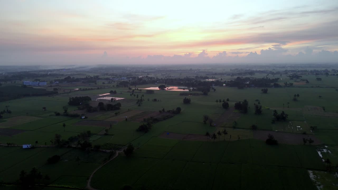 Aerial view of a captures vast, peaceful green farmlands at Sunrise. Reflective fields, scattered trees, and distant homes create a tranquil, natural landscape. Ideal for rural themes