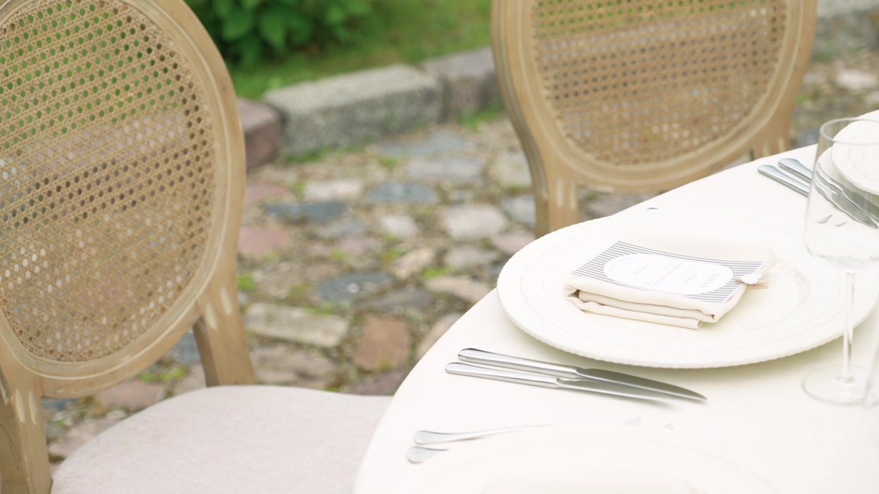 Bright outdoor scene featuring a stylish white table setup with stacked napkins, plates, glassware, cutlery, vintage wooden chairs, and cobblestone patio under soft natural light.