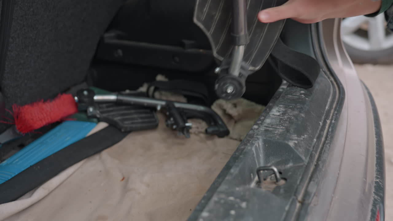 Car Trunk Open With Wheelchair Parts, Hand Reaching For Frame And Tools, Dusty Bumper And Liner, Folded Ramp And Securing Straps, Practical Preparation For Accessible Travel, Roadside Scene