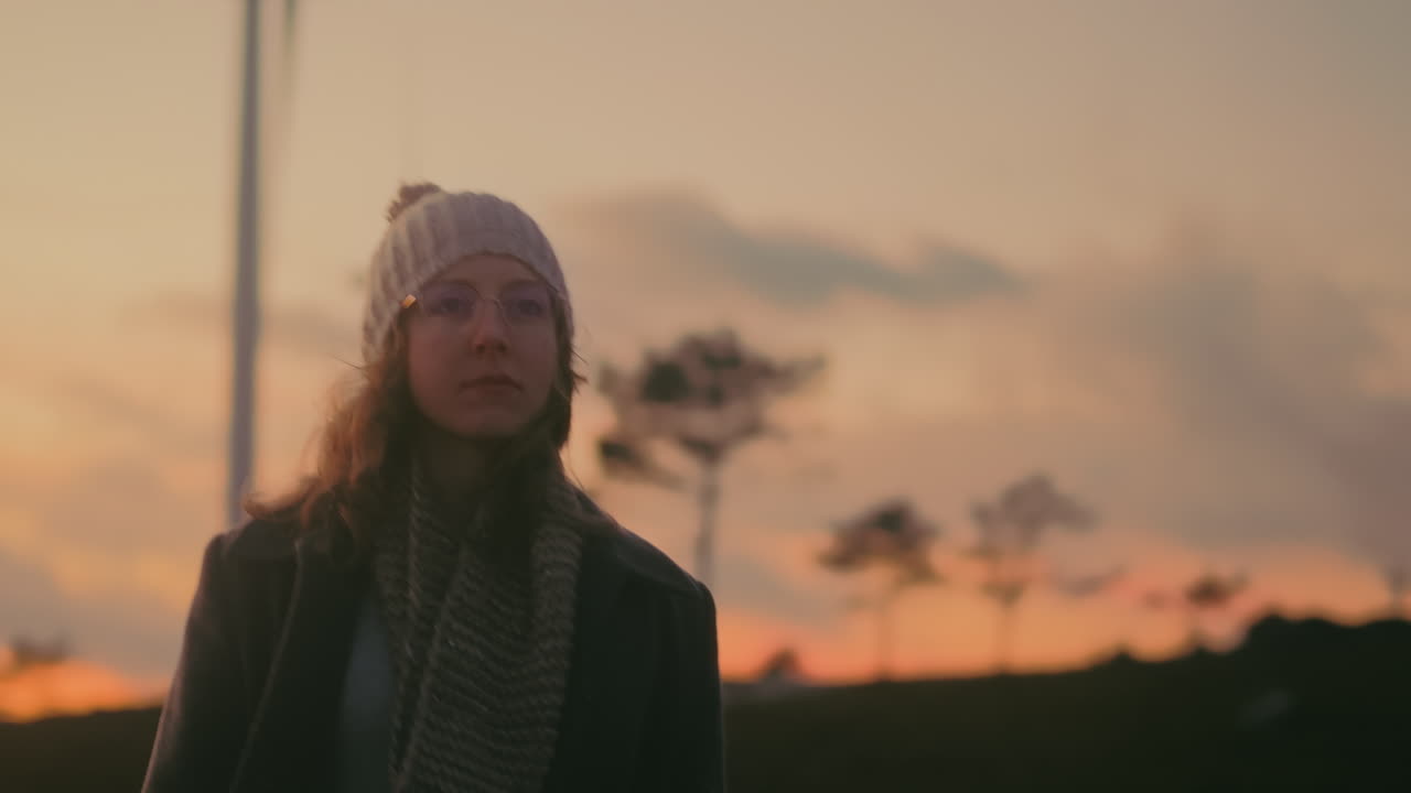 Young Caucasian woman with blonde hair, glasses, beanie, gray coat, and scarf walks on a mountain road in Portugal at sunset. She enjoys the scenic landscape and renewable energy wind turbines.