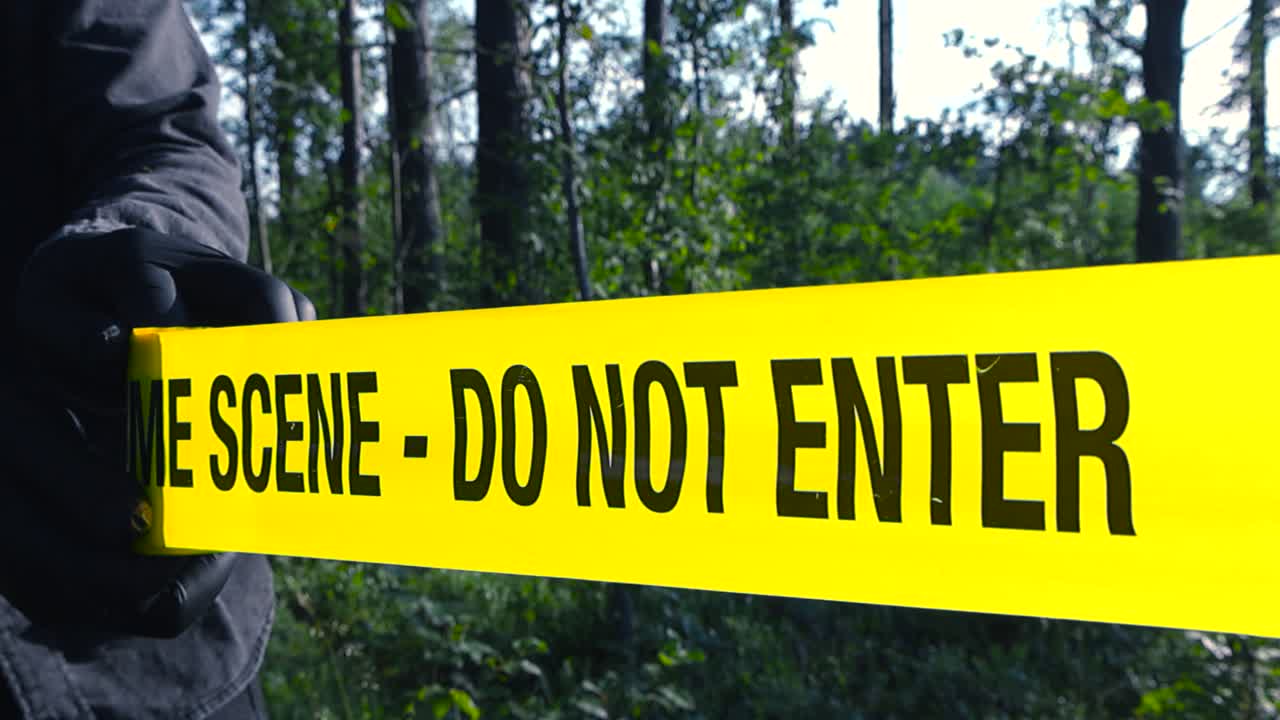 Yellow crime scene tape in close up being pulled in front of blurry wooded area with bushes. Forensic detective with black gloves, hands unrolling barricade tape, marking scene during sunny daylight