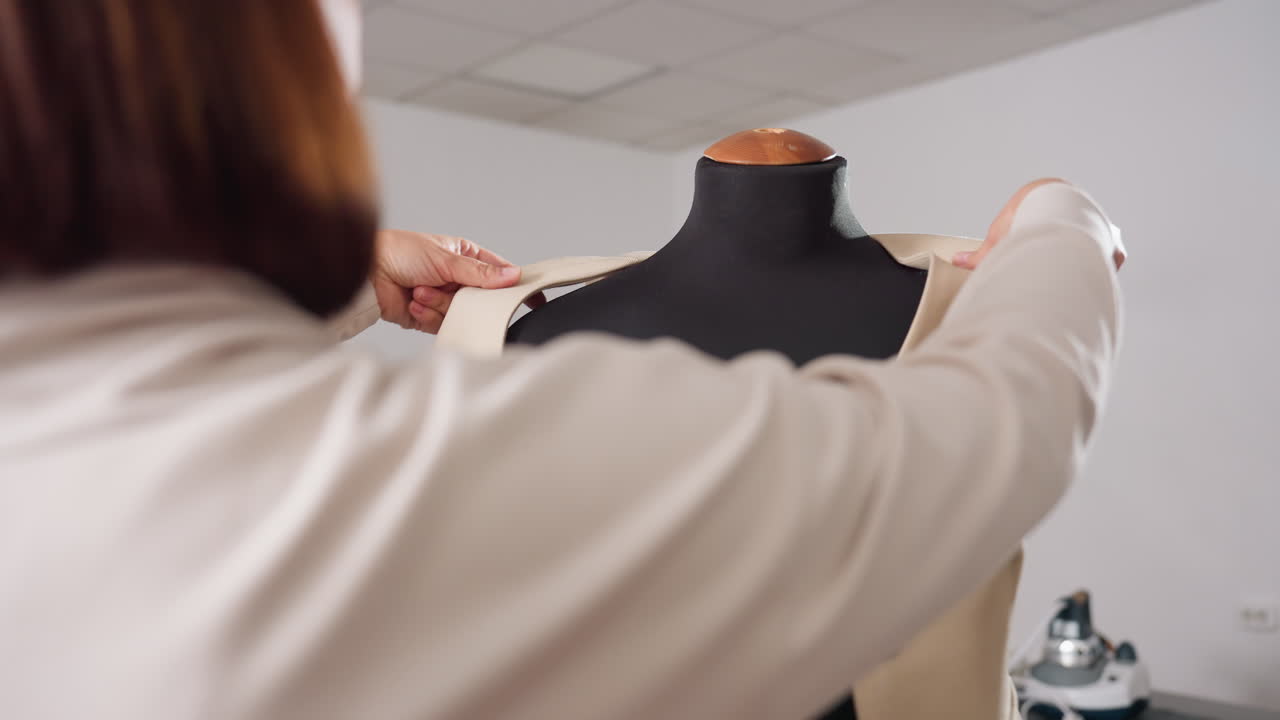 Clothing designer lifts beige sleeveless blazer from black dress form in workshop, sliding fingers over seams, checking fit before next tailoring step, highlighting precision garment handling
