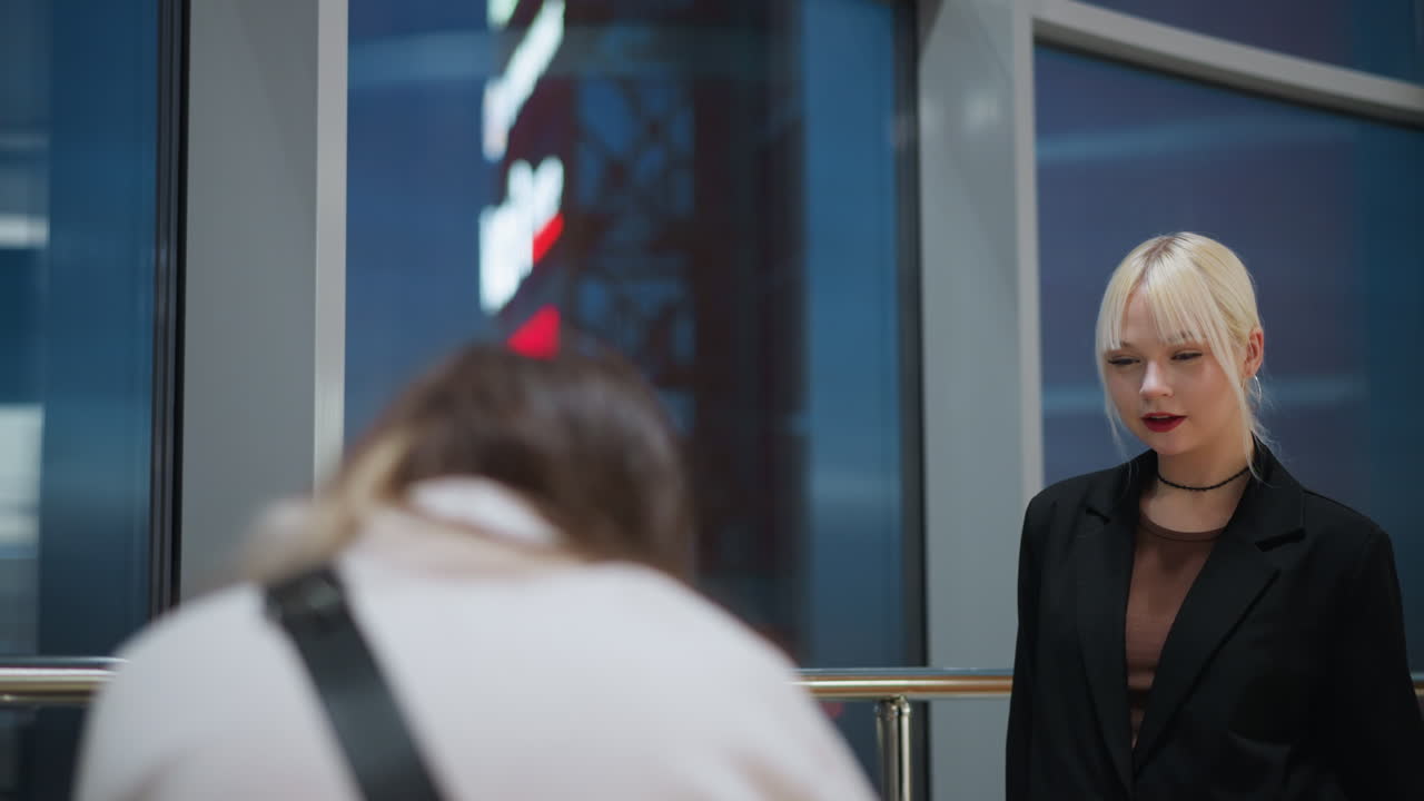 Elegant blonde girl in black jacket posing indoors with playful facial expression while being photographed near glass window as colorful city lights glow outside in modern shopping mall