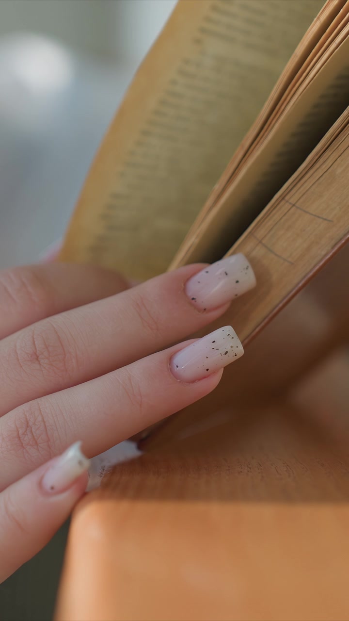 Close up of young woman hands with light pink nail polish. Focus remains on fingers interacting with yellowed book pages. Soft indoor lighting creates gentle shadows