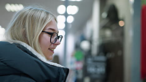 young girl in glasses and black hoodie walking through well-lit mall, looking happy, blurred background features retail space with people passing by, showcasing modern shopping environment