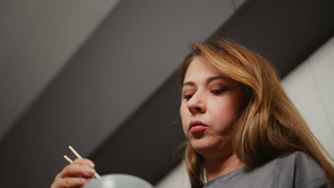 upward angle captures woman in ash top lifting head while chewing pasta with chopsticks in modern kitchen, under sleek cabinetry with soft lighting ambiance