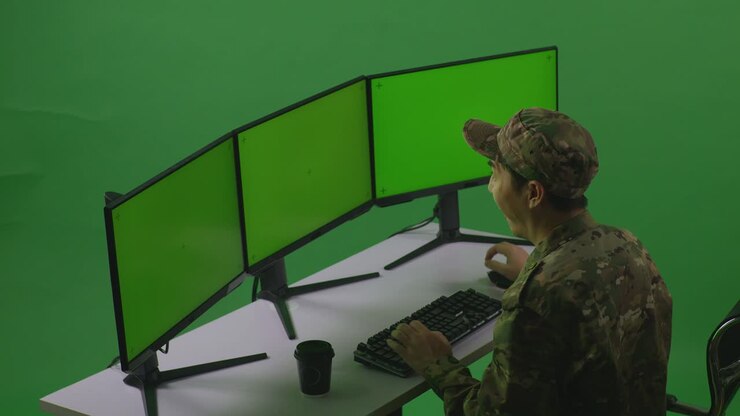 Sidebillede af en glad asiatisk mandlig soldat, der fejrer, mens han arbejder med flere computerskærme med mockup i et green screen-studie.