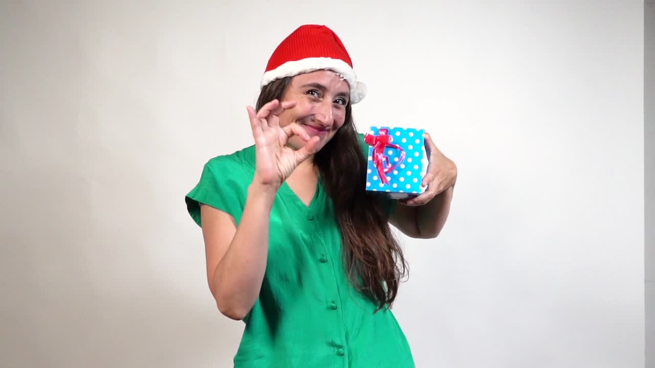 Latin female in Santa hat and green blouse signals OK while presenting blue polka-dot gift