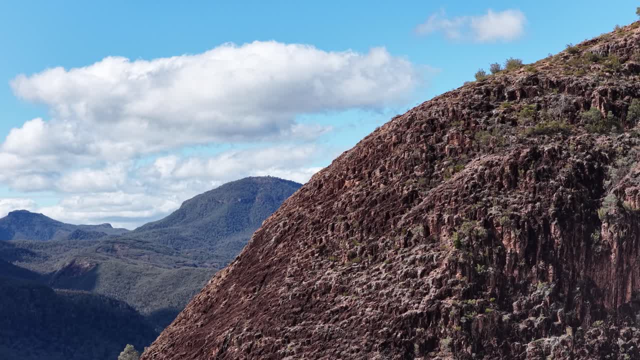Camera slowly pans right across a rugged, sunlit cliff face with distant mountain ranges and blue sky, highlighting natural textures and dramatic landscape