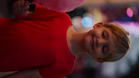 Vertical-video-portrait-of-a-happy-little-blond-boy-with-blue-hair-in-a-red-T-shirt-who-looks-appetizingly-at-pink-cotton-candy-and-bites-it-in-the-amusement-park