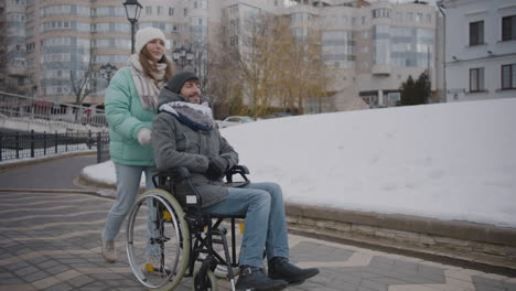 Glückliche-Kaukasische-Frau,-Die-Ihren-Behinderten-Freund-Im-Rollstuhl-Für-Einen-Spaziergang-In-Der-Stadt-Nimmt-Und-Miteinander-Redet