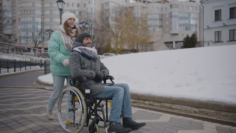 Happy-Caucasian-woman-taking-her-disabled-friend-in-wheelchair-for-a-walk-in-the-city-and-talking-together