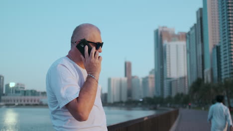 Happy-handsome-senior-man-in-sunglasses-and-white-t-shirt-talking-on-the-phone-standing-on-the-waterfront-in-summer-against-the-city-and-buildings