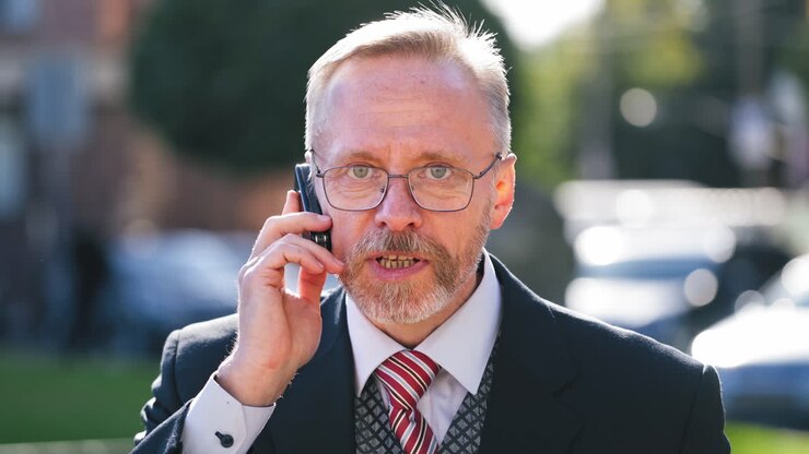 Serious businessman wearing eyeglasses talks on phone while looking at camera. Portrait of a senior man in suit and tie talking on a cell phone in the city.