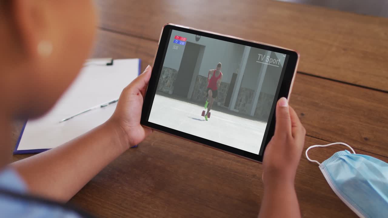 태블릿에서 육상 경기를 시청하는 얼굴 마스크와 함께 테이블에 앉아있는 여성의 복합