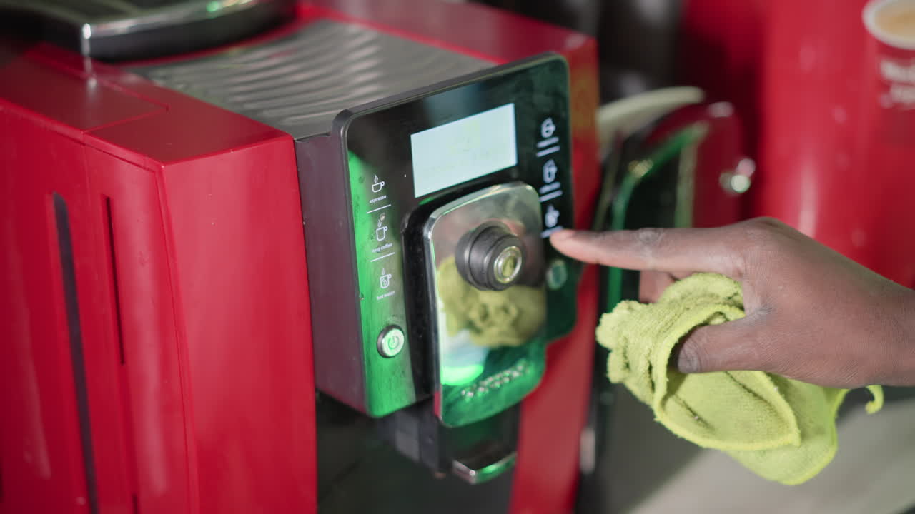 Close-up of hand cleaning red coffee machine control panel with green cloth, focusing on wiping down surface and ensuring machine is spotless and well-maintained