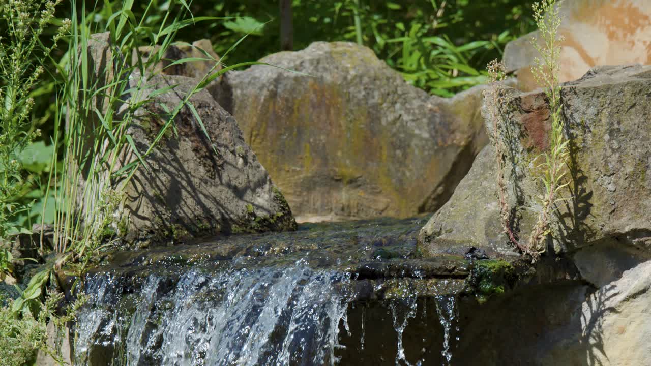 Small waterfall cascades over natural rocks in a lush Berlin park. Bright daylight, steady camera, and vibrant greenery create a peaceful outdoor atmosphere