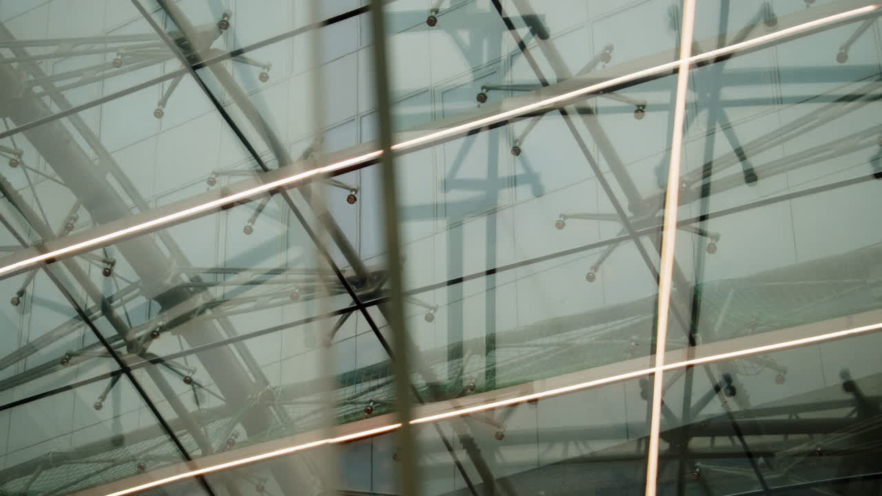 Abstract view of a modern glass roof with intersecting steel beams and transparent panels. Geometric lines and reflections create a minimal, contemporary architectural background. Suitable for concept