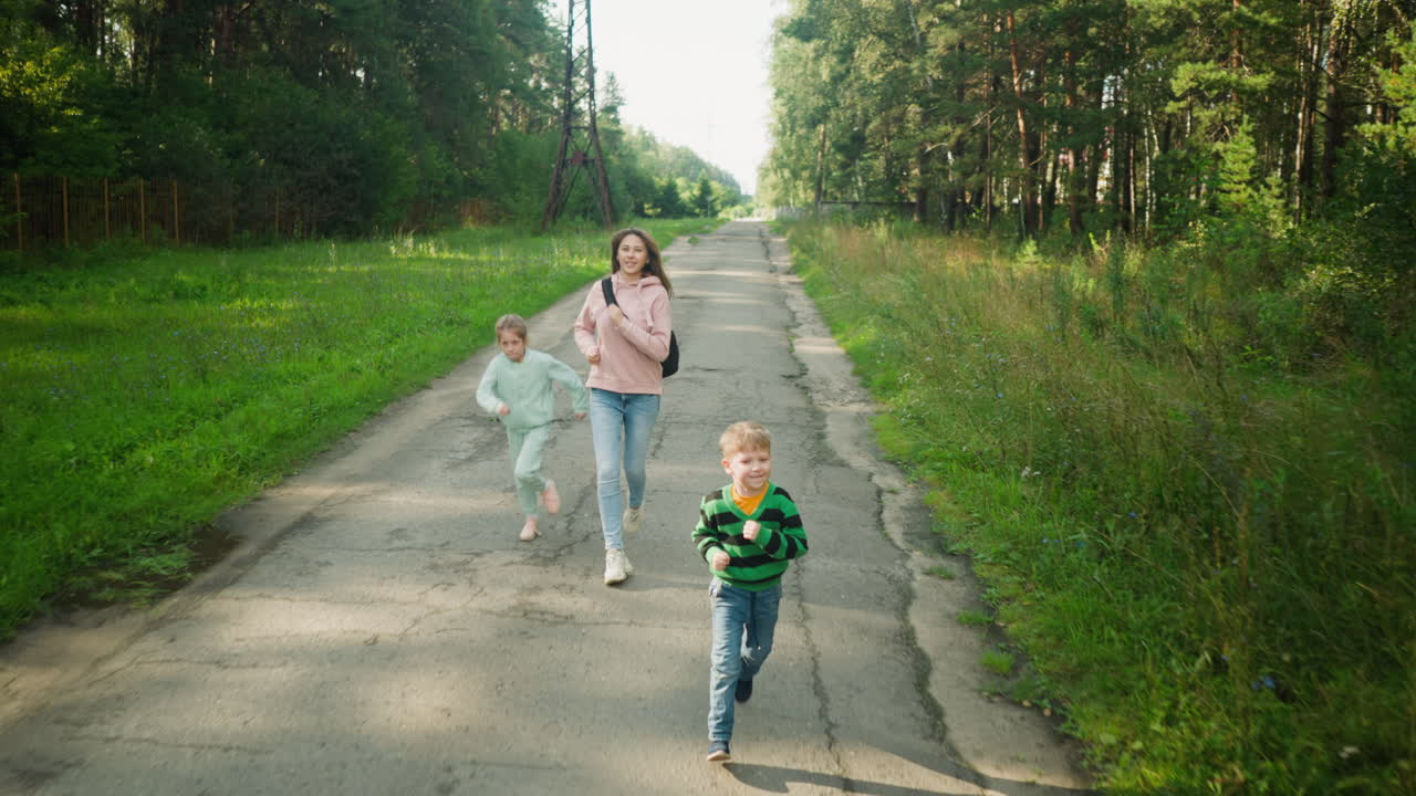 Smiling young boy runs ahead joyfully while mother and sister follow behind on cracked tarred countryside road, surrounded by lush green grass and forest trees during daylight