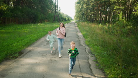 smiling young boy runs ahead joyfully while mother and sister follow behind on cracked tarred countryside road, surrounded by lush green grass and forest trees during daylight