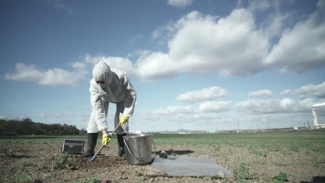 científico en traje protector excavando arcilla de campo contaminada, fondo industrial, concepto de desastre de catástrofe ecológica