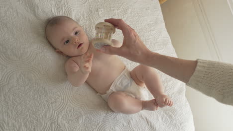 mother gives happy baby toy as his laying on his back playing in white bed sheets, bright bedroom