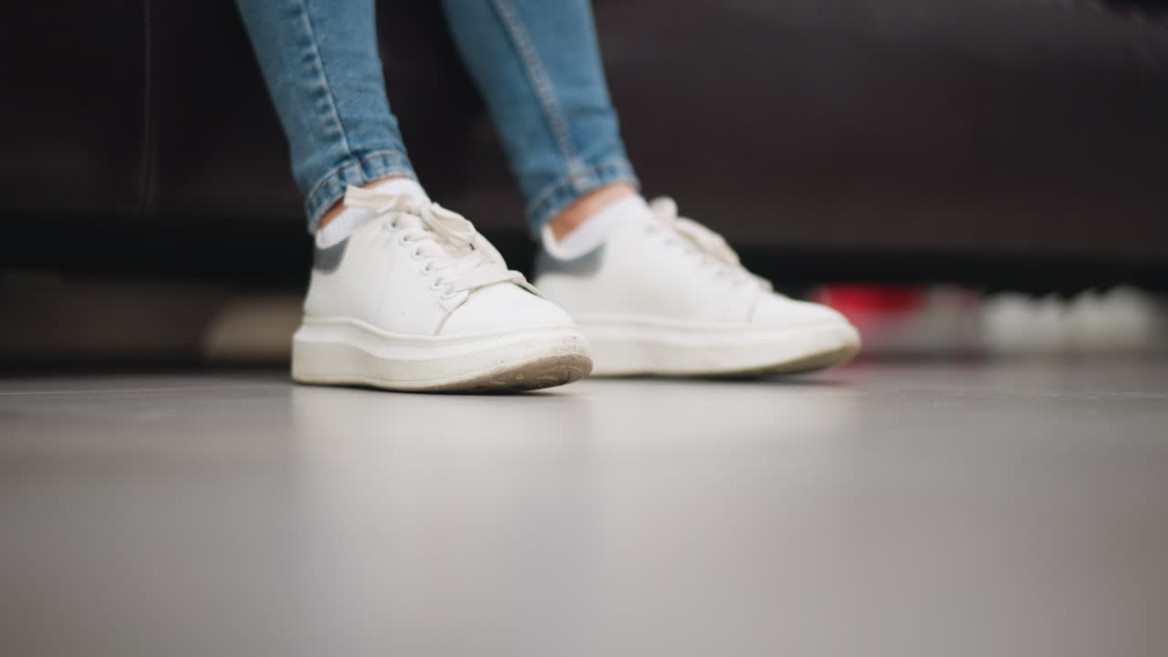 여성의  캔버스 스니커의 클로즈업은 편안한 쇼핑몰 설정에서 소파에 앉아있는 동안 청바지 바지와 함께 바닥에 왼쪽과 오른쪽 발을 부드럽게 합니다.