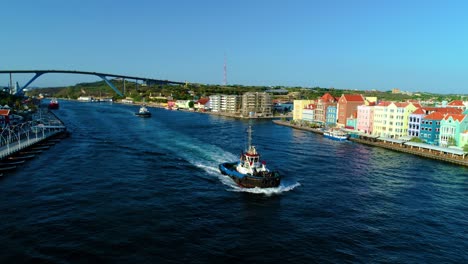 Pair-of-tug-boats-drive-along-bay-entrance-in-Willemstad,-Curacao,-Queen-Juliana-Bridge-behind