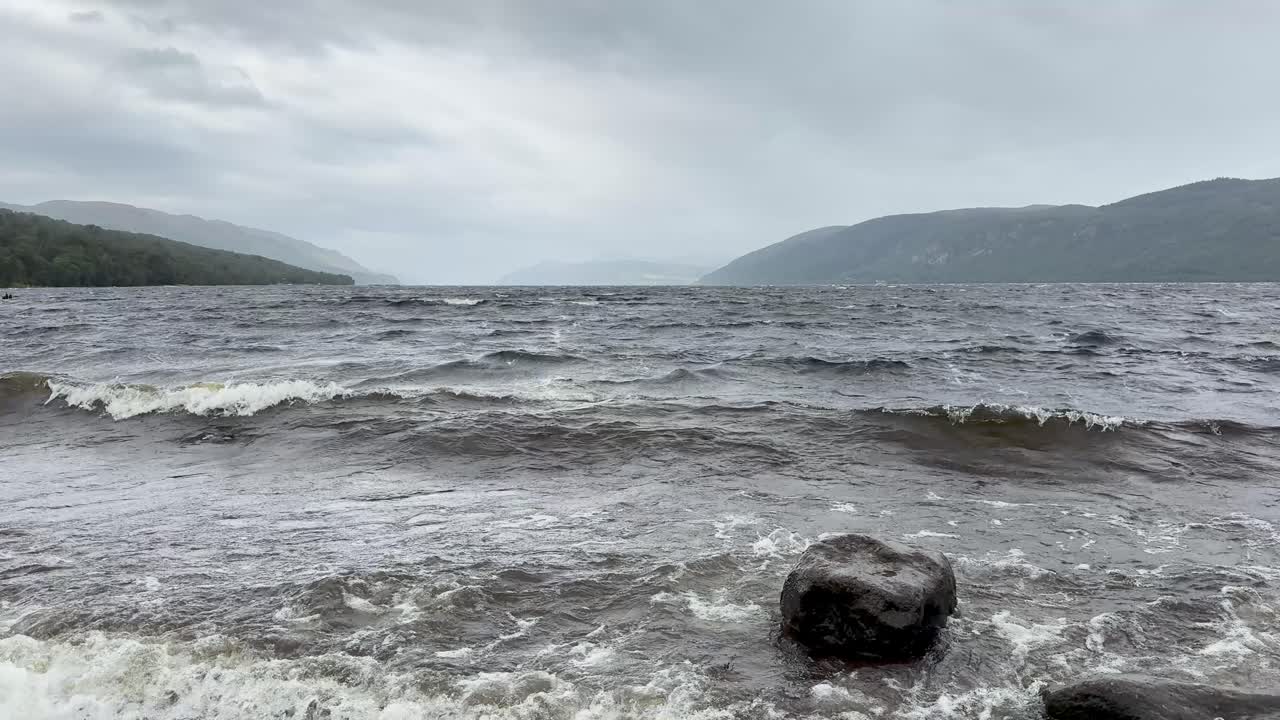 Choppy waves hit rocky Loch Ness shore under overcast skies, static camera, natural daylight