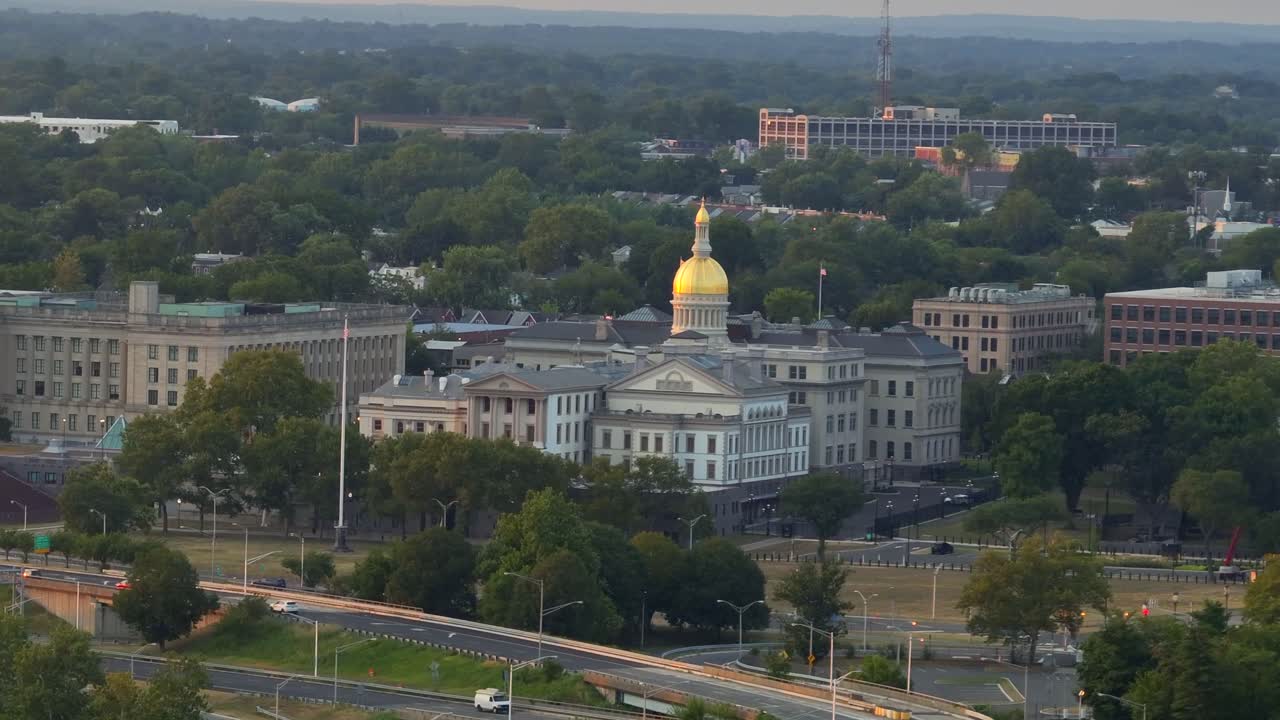 Aerial view of New Jersey State House in Trenton at sunset time in New Jersey. Wide shot. Waving American flag on flagpole. Cars on road along Delaware River