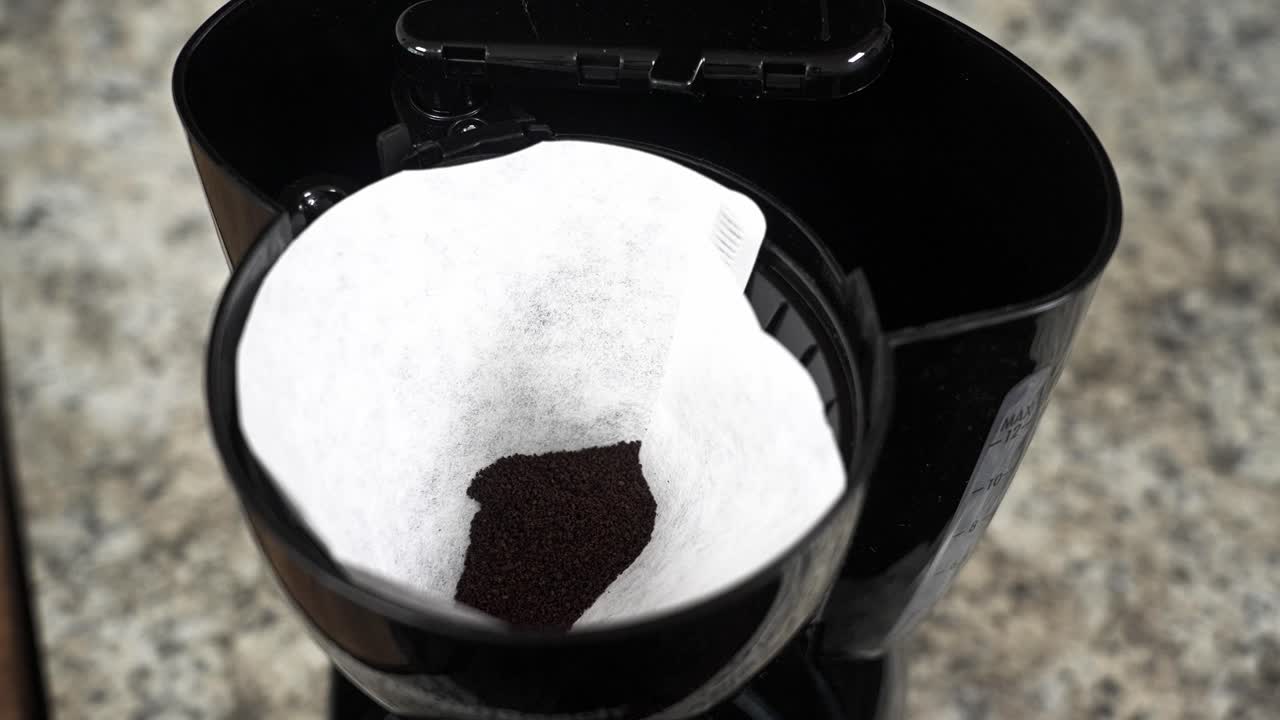 Close-up top view of hands placing a paper filter and spooning fresh ground coffee into a drip coffee maker, preparing a morning brew