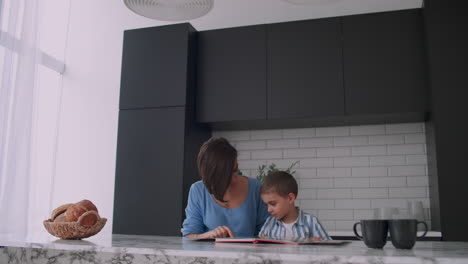 joven madre y hijo españoles sentados a la mesa leyendo un libro juntos y sonriendo