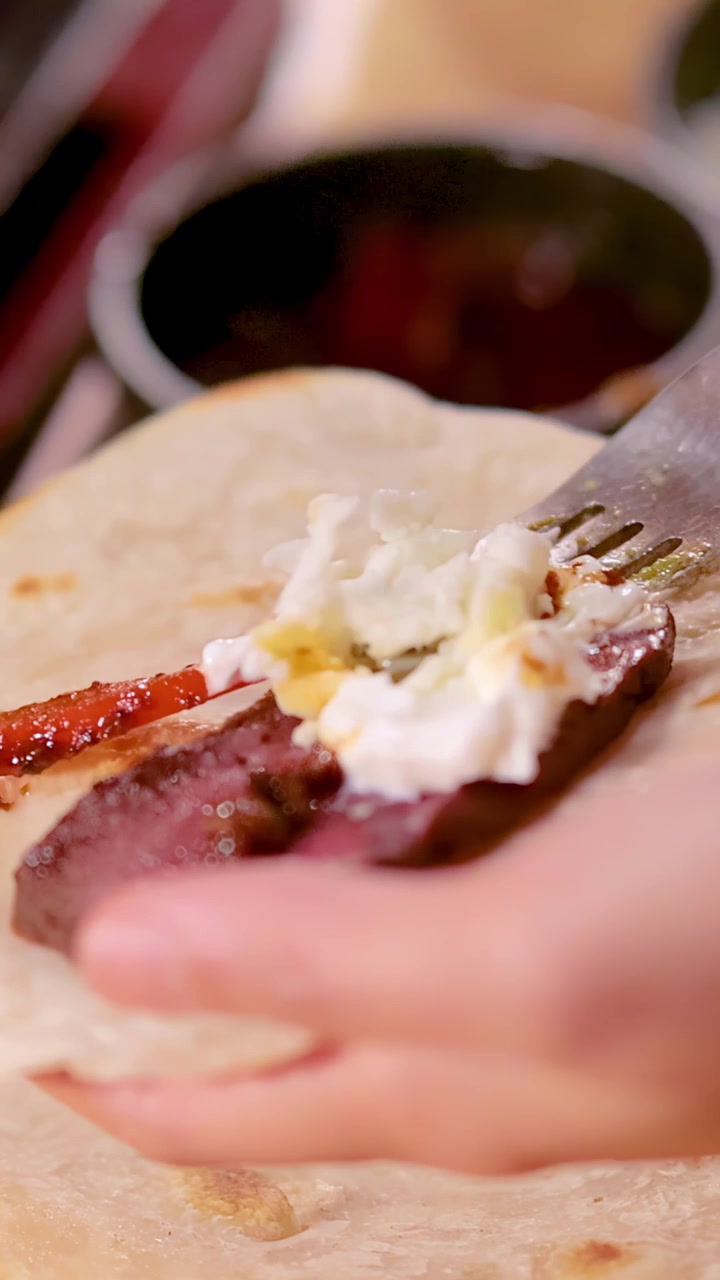 Una mano aggiunge una salsa cremosa a un taco di manzo su tortilla sotto una luce calda e soffusa