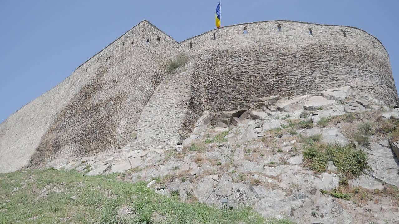 Mountain cliff with rough rock face and sparse vegetation under bright sky, tilted up to fort built above