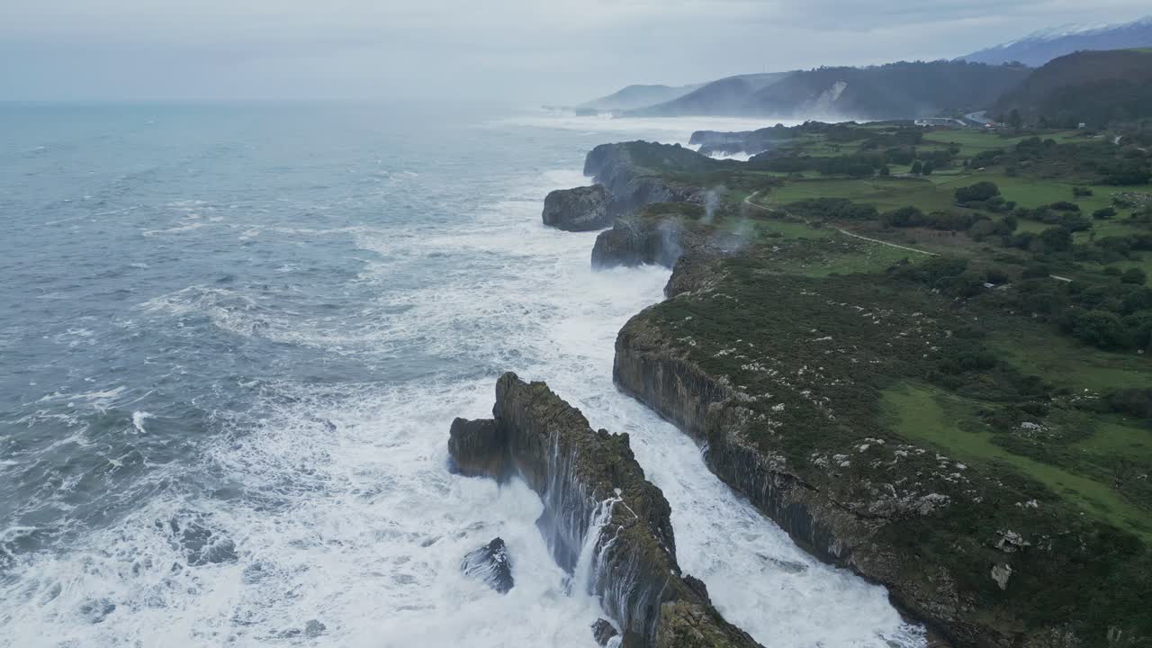 Massive waves crash against a rugged cliffside coastline, flowing through a natural rock arch. In the background, towering mountains create a dramatic scene, showcasing the raw power of the ocean.