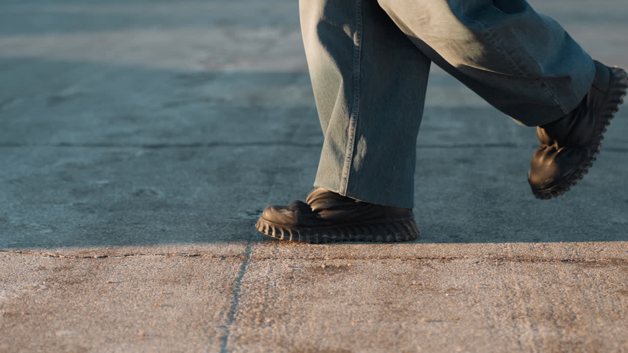 Piedi in primo piano che camminano su un marciapiede illuminato dal sole, stivali decisi che toccano terra, pantaloni di jeans che ondeggiano, lunghe ombre che si allungano, passo svelto e ritmo sicuro, atmosfera di pendolarismo urbano, testurizzato.