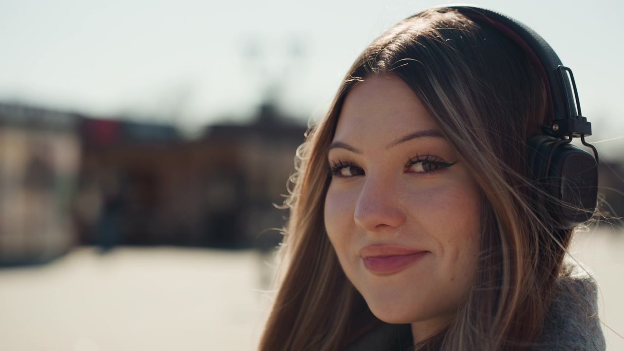 Donna casual con cuffie che si gode la scena cittadina. Allegra donna asiatica in cappotto grigio che ascolta musica all'aperto. Donna influente con sguardo giocoso e sorriso caloroso in un ambiente urbano all'aperto