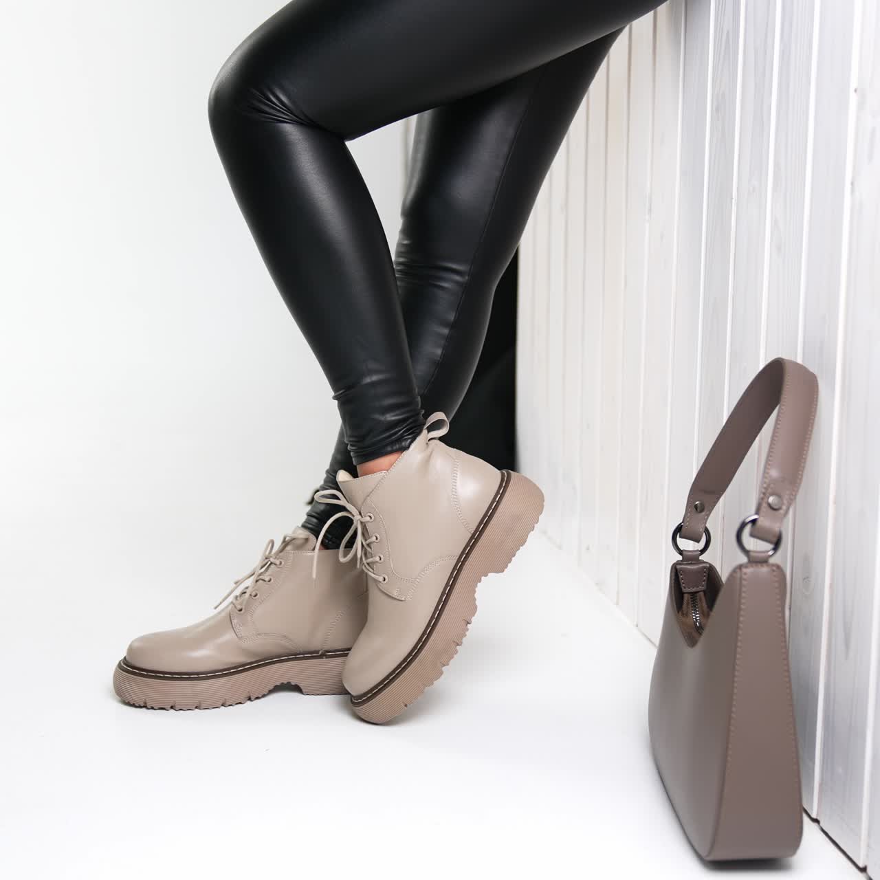 Woman in black leather pants and beige boots standing at the wall. Beige bag beside at the wall. White backdrop