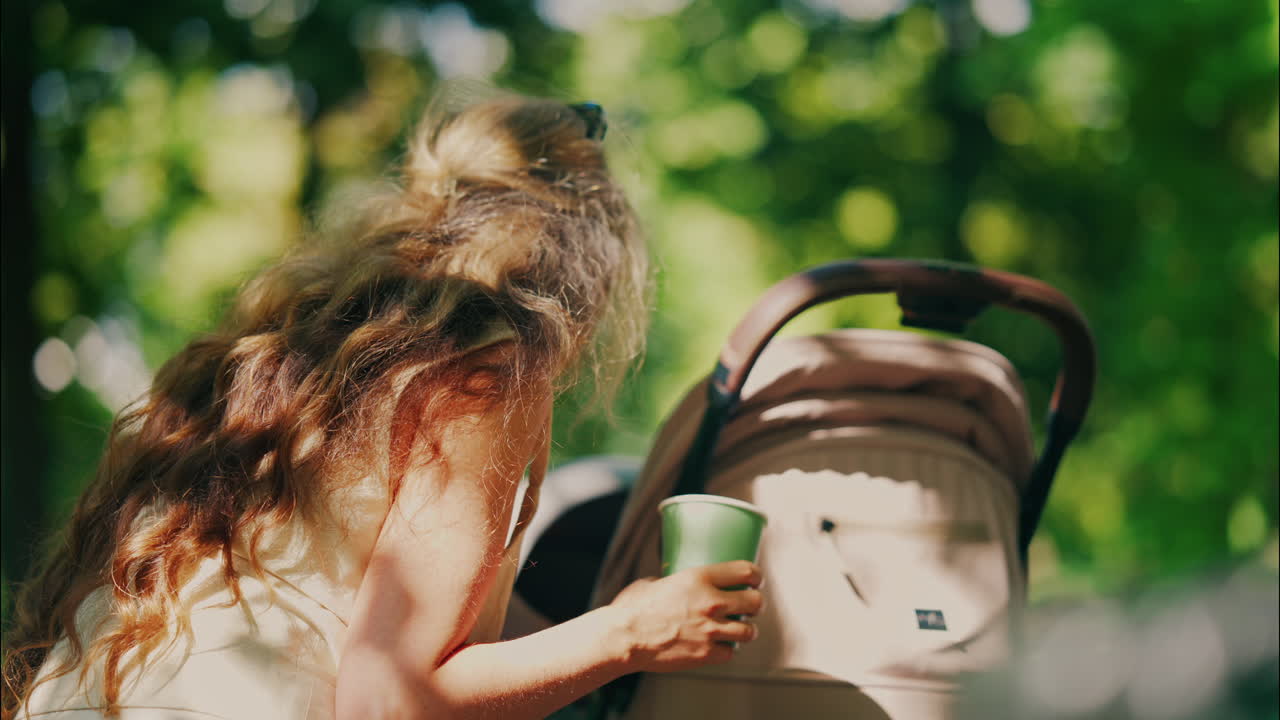 Ung mor med langt krøllet hår holder en kaffekop, mens hun læner sig over en barnevogn i parken