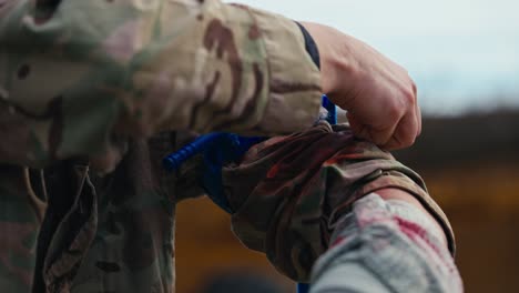 close-up of a confident male soldier in a green camouflage uniform puts a blue tourniquet on his arm to avoid blood loss when wounded during first aid training at a military training ground