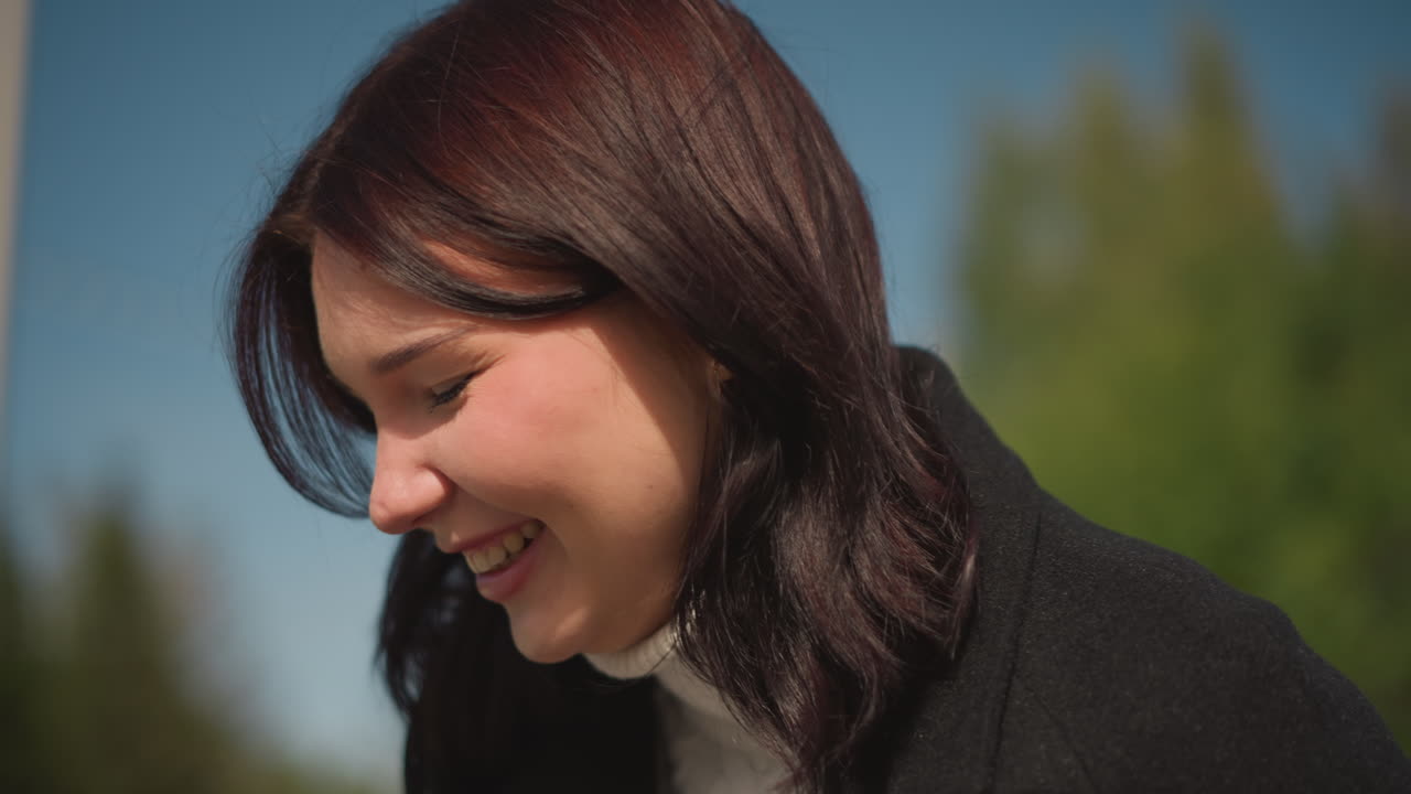 검은 코트를 입은 여성이 머리카락을 조정하고 드폰을 들고 따뜻하게 미소 짓는 클로즈업, 야외에서 이 많은 공원, 배경에는 활기찬 초록색