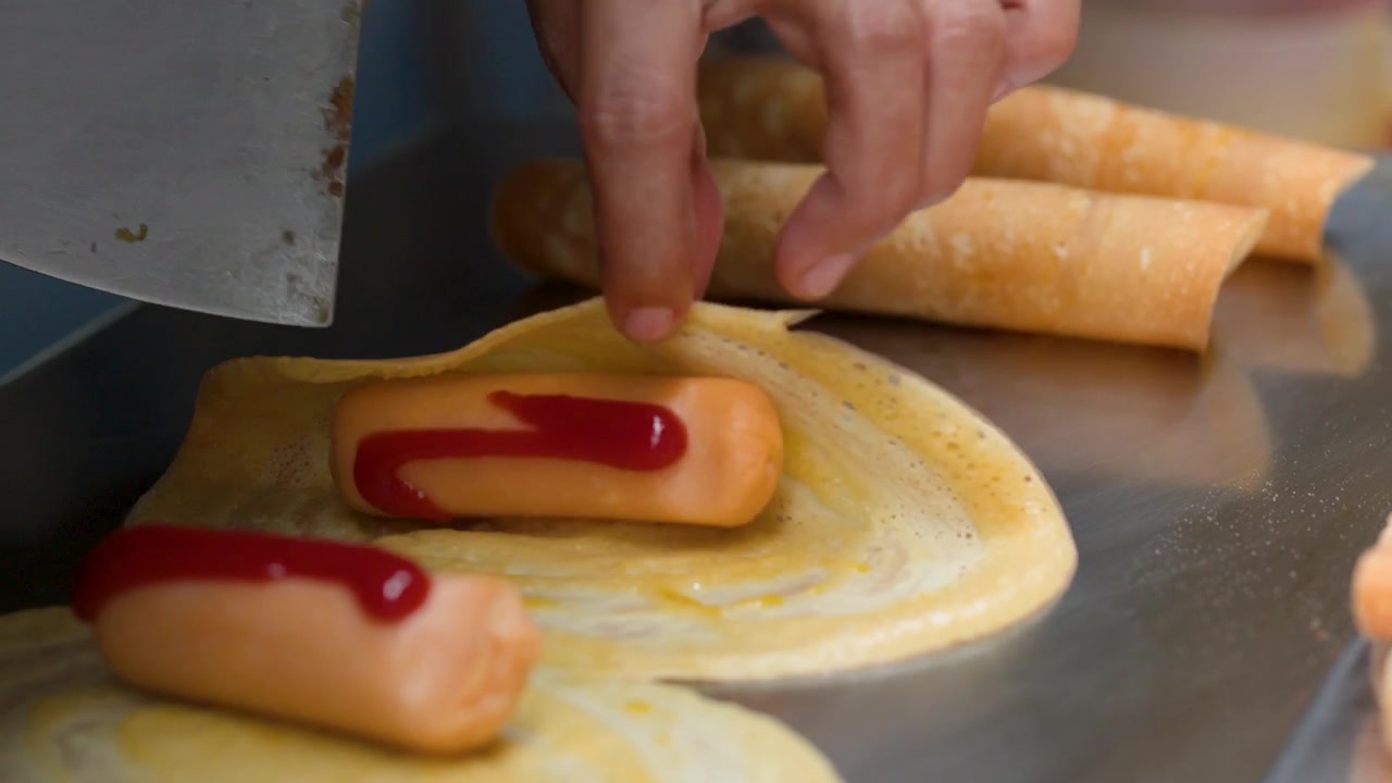 Primo piano di mani che arrotolano frittelle sottili con salsicce e ketchup su una piastra di metallo.