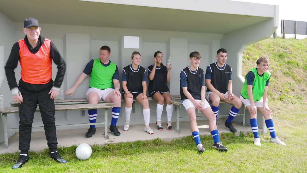 Male and female soccer players sitting on bench and supporting with coach observing game on pitch