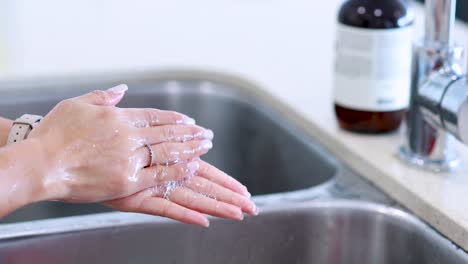 a person washes hands with soap under running water at a stainless steel kitchen sink, using proper technique. bright, natural lighting, steady camera